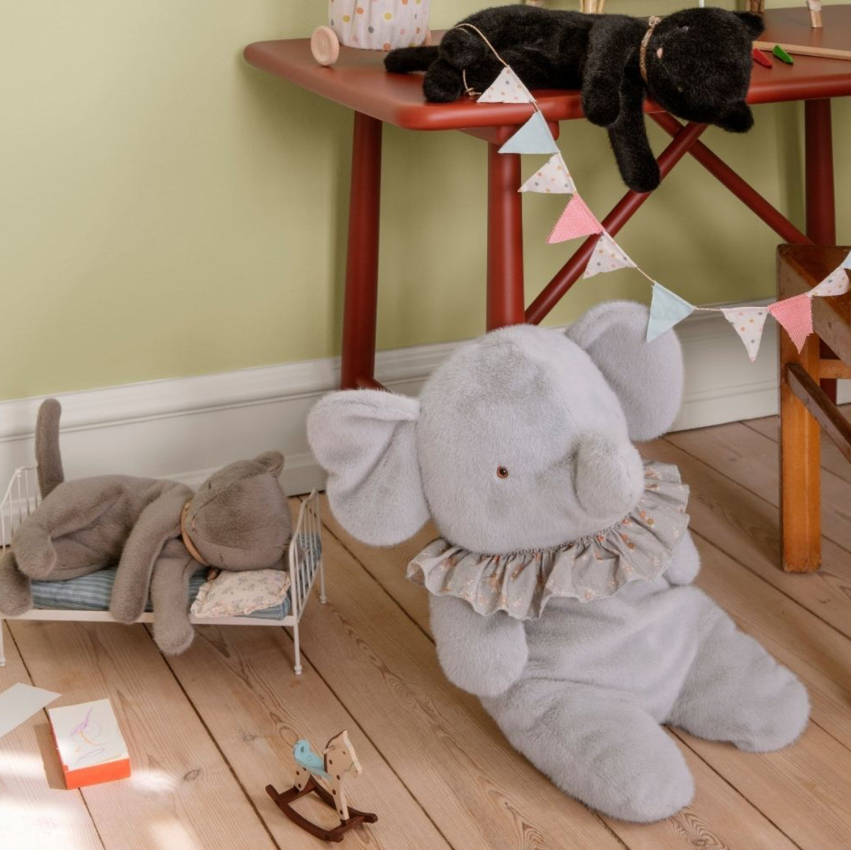 Gray elephant plush toy on a wooden floor with a child's play area in the background.